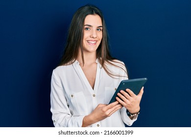Young brunette teenager using touchpad device looking positive and happy standing and smiling with a confident smile showing teeth 