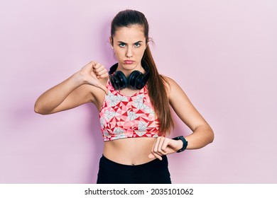 Young Brunette Teenager Wearing Gym Clothes, Using Headphones And Smart Watch With Angry Face, Negative Sign Showing Dislike With Thumbs Down, Rejection Concept 