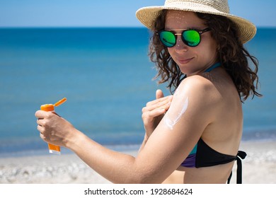Young brunette woman applying suntan solar cream to her shoulder from a plastic container, wearing sunglasses and summer hat with ocean in background, Sunscreen spf lotion. Copy space.