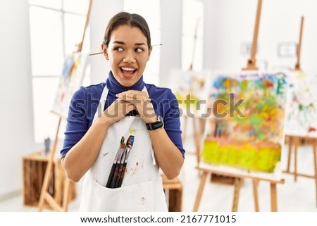 Young brunette woman at art studio laughing nervous and excited with hands on chin looking to the side 
