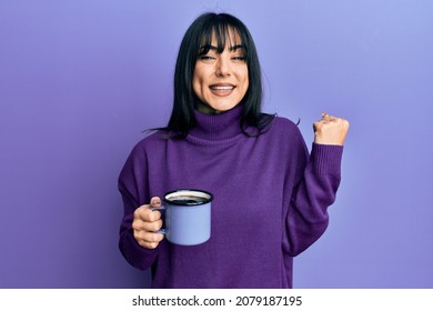 Young brunette woman with bangs drinking a cup of coffee screaming proud, celebrating victory and success very excited with raised arm 
