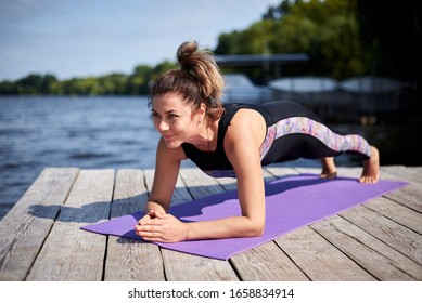 Young Brunette Woman With Bare Feet, Wearing Black And Purple Fitness Outfit, Doing Plank On Violet Yoga Mat On Wooden Pier In Summer. Power Training Outside By The Lake. Healthy Lifestyle Concept.