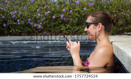 Young brunette woman in bikini and sunglasses types on smartphone sitting in swimming pool side view
