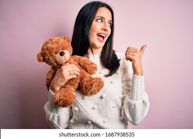 Young brunette woman with blue eyes hugging teddy bear stuffed animal over pink background pointing and showing with thumb up to the side with happy face smiling