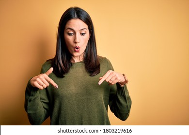 Young brunette woman with blue eyes wearing green casual sweater over yellow background Pointing down with fingers showing advertisement, surprised face and open mouth
