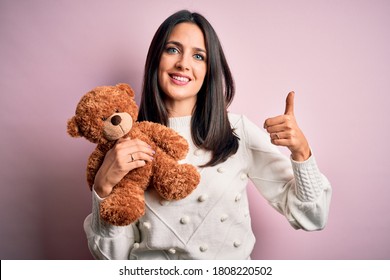 Young brunette woman with blue eyes hugging teddy bear stuffed animal over pink background happy with big smile doing ok sign, thumb up with fingers, excellent sign
