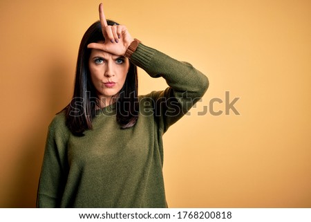 Young brunette woman with blue eyes wearing green casual sweater over yellow background making fun of people with fingers on forehead doing loser gesture mocking and insulting.