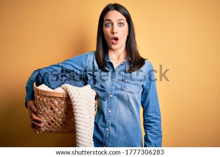 Young brunette woman with blue eyes doing domestic chores holding laundry basket scared in shock with a surprise face, afraid and excited with fear expression