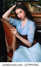 Young brunette woman in blue vintage dress and corset sitting on old chair in old fashioned room