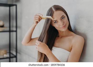 Young Brunette Woman Brushes Hair With Comb After Taking Shower And Applying Hair Care Mask, Wears Minimal Makeup, Has Healthy Glowing Skin After Hygienic Procedures, Stands Wrapped In Towel