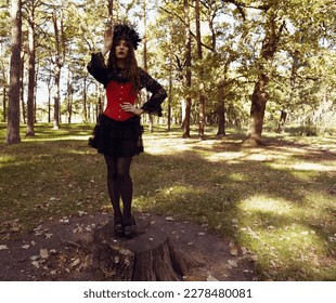 A young brunette woman in a carnival costume stands on a stump in the forest. Halloween woman portrait. Woman in dark suit - Shutterstock ID 2278480081