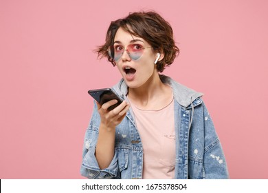 Young Brunette Woman In Casual Denim Jacket, Eyeglasses Posing Isolated On Pastel Pink Background Studio Portrait. People Lifestyle Concept. Mock Up Copy Space. Talking On Mobile Phone With Air Pods