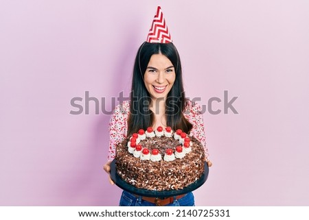 Young brunette woman celebrating birthday holding big chocolate cake smiling and laughing hard out loud because funny crazy joke. 