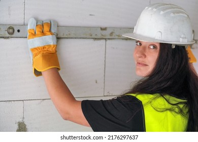 Young Brunette Woman Construction Worker In White Hard Hat And Green High Visibility Vest, Measuring Wall With Spirit Bubble Level, Looking Back Over Her Shoulder