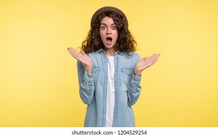 Young brunette woman with curly hair opened her mouth in a surprise, isolated over yellow background
