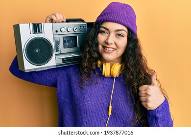Young brunette woman with curly hair holding boombox, listening to music smiling happy and positive, thumb up doing excellent and approval sign  - Shutterstock ID 1937877016