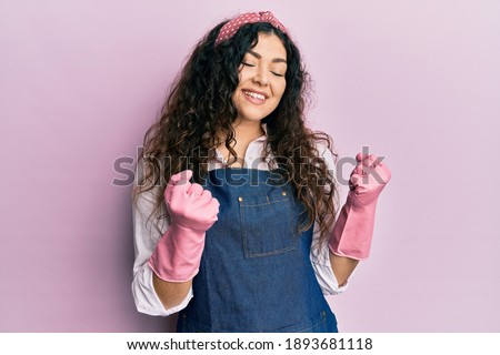 Young brunette woman with curly hair wearing cleaner apron and gloves excited for success with arms raised and eyes closed celebrating victory smiling. winner concept. 