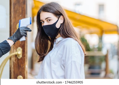 Young brunette woman customer in facial mask coming in a cafe and getting her temperature measured from forehead with infrared thermometer. Concept of a new social rules after coronavirus pandemic. - Powered by Shutterstock