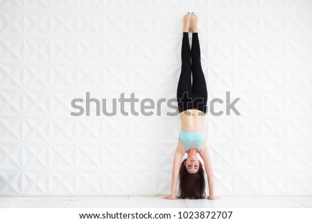 Young brunette woman doing a handstand over a white wall with copy space. Slim sporty woman practicing balance pose