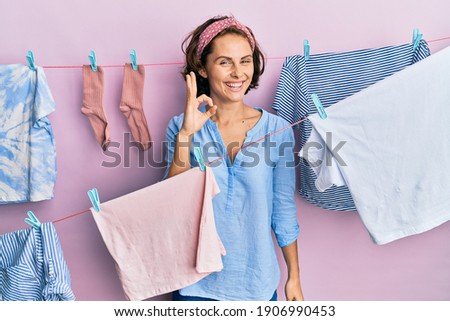 Young brunette woman doing laundry around string hangs smiling positive doing ok sign with hand and fingers. successful expression. 