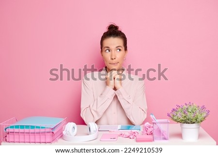 Young brunette woman dreaming at the work desk, she bite her lip and looks a side, isolated next to pink wall