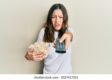 Young brunette woman eating popcorn using tv control clueless and confused expression. doubt concept.  - Powered by Shutterstock