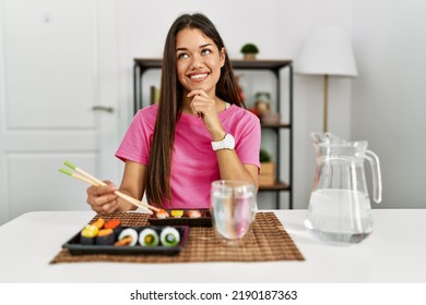 Young Brunette Woman Eating Sushi Using Chopsticks With Hand On Chin Thinking About Question, Pensive Expression. Smiling And Thoughtful Face. Doubt Concept. 