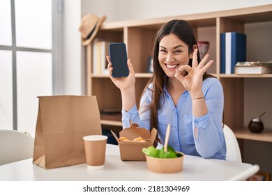 Young Brunette Woman Eating Take Away Food At Home Showing Smartphone Screen Doing Ok Sign With Fingers, Smiling Friendly Gesturing Excellent Symbol 