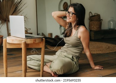 Young brunette woman in eyeglasses at home sitting on the floor using laptop and listening to music