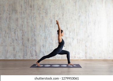 Young Brunette Woman Fitness Instructor Wearing A Black One-piece Suit Doing Virabhadrasana Yoga Standing On A Mat On A Wooden Floor Against A Background Of A Concrete Wall