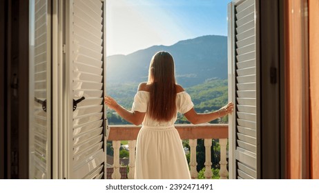 Young brunette woman in a flowing dress opens balcony doors, steps onto the terrace, and revels in a breathtaking mountain sunrise or sunset - Powered by Shutterstock