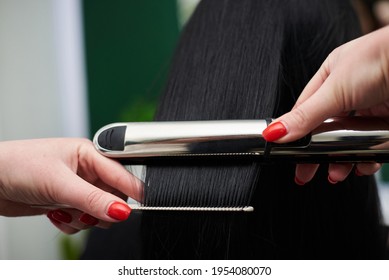 Young Brunette Woman Getting Ready For Party. Professional Hairdresser Making Hairstyling For Female Client With Hair Straightener In Front Of Mirror. Close-up Picture Of Work Process In Barber Shop.