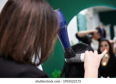 Young Brunette Woman Getting Ready For Party. Professional Hairdresser Making Hairstyling For Female Client With Hair Dryer In Front Of Big Mirror. Close-up Picture Of Work Process In Barber Shop.