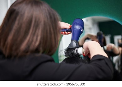 Young Brunette Woman Getting Ready For Party. Professional Hairdresser Making Hairstyling For Female Client With Hair Dryer In Front Of Big Mirror. Close-up Picture Of Work Process In Barber Shop.