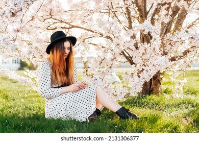 Young brunette woman in hat and dress sitting near white pink blooming tree. Generation Z girl enjoy spring mood during a sunny day. Relax on the nature. Springtime blossom. Selective focus.