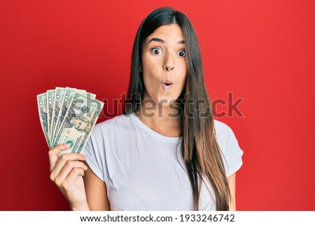 Young brunette woman holding 20 dollars banknote scared and amazed with open mouth for surprise, disbelief face 