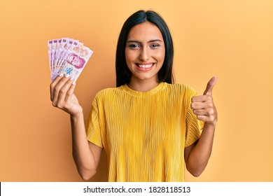 Young brunette woman holding 50 mexican pesos banknotes smiling happy and positive, thumb up doing excellent and approval sign 