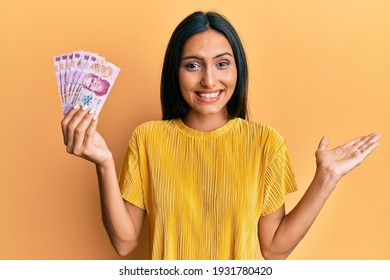 Young brunette woman holding 50 mexican pesos banknotes celebrating achievement with happy smile and winner expression with raised hand 