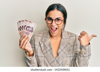Young brunette woman holding 500 mexican pesos banknotes pointing thumb up to the side smiling happy with open mouth 