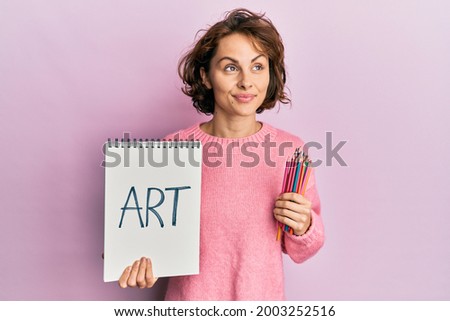 Young brunette woman holding art notebook and colored pencils smiling looking to the side and staring away thinking. 