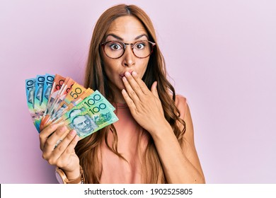 Young brunette woman holding australian dollars covering mouth with hand, shocked and afraid for mistake. surprised expression 