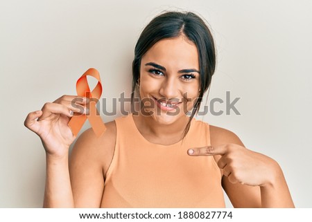 Young brunette woman holding awareness orange ribbon pointing finger to one self smiling happy and proud 