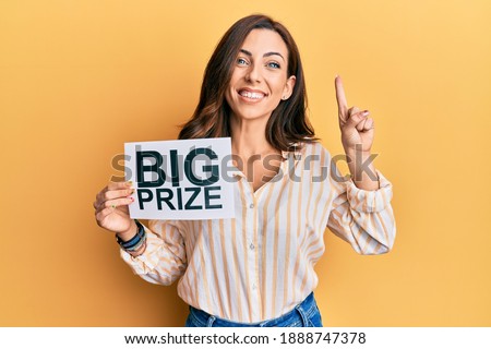 Young brunette woman holding big prize paper smiling with an idea or question pointing finger with happy face, number one 