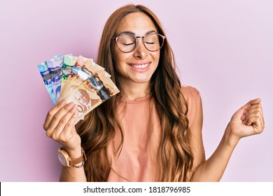 Young brunette woman holding canadian dollars screaming proud, celebrating victory and success very excited with raised arm 