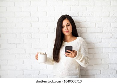 Young brunette woman holding a cup of marshmallow cocoa and using her mobile phone on white brick wall background - Powered by Shutterstock