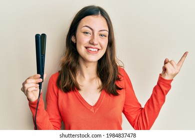 Young Brunette Woman Holding Hair Straightener Smiling Happy Pointing With Hand And Finger To The Side 