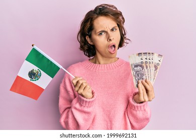 Young brunette woman holding mexico flag and mexican pesos banknotes in shock face, looking skeptical and sarcastic, surprised with open mouth 