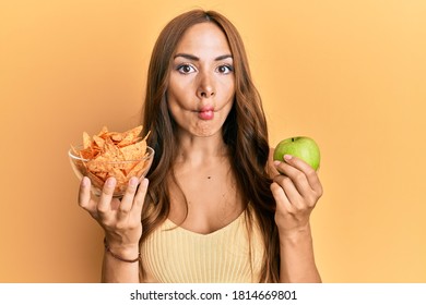 Young Brunette Woman Holding Nachos And Healthy Green Apple Making Fish Face With Mouth And Squinting Eyes, Crazy And Comical. 