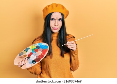 Young brunette woman holding paintbrush and palette wearing beret skeptic and nervous, frowning upset because of problem. negative person. 