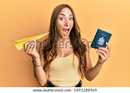 Young brunette woman holding paper airplane and canadian passport celebrating crazy and amazed for success with open eyes screaming excited. 